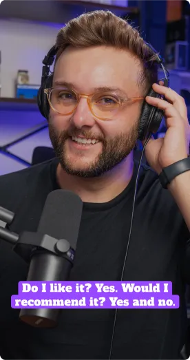 Smiling man with glasses and beard wearing headphones and speaking into a microphone in a recording studio.